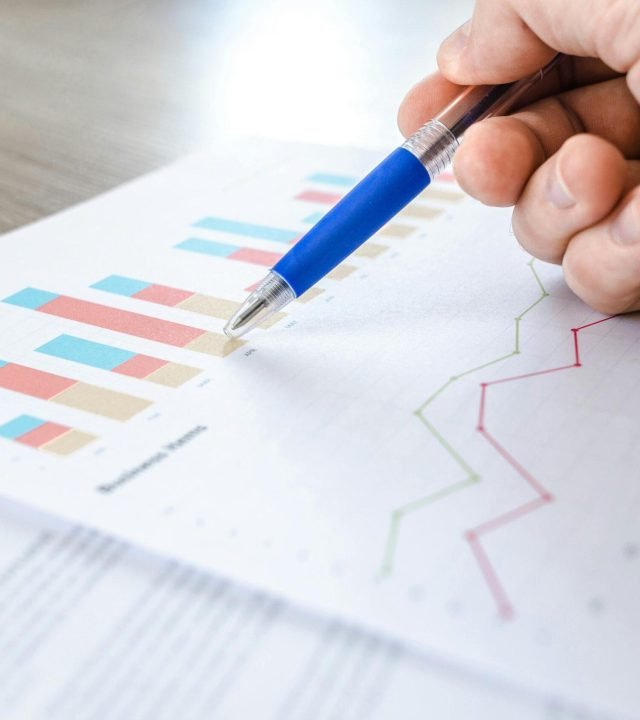 A close-up of a hand with a pen analyzing data on colorful bar and line charts on paper.
